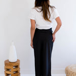 Woman wearing a white t-shirt with text and graphics, black satin maxi skirt, and beige hat in a minimalistic setting. Made in Canada Back View.