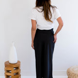 Woman wearing a white t-shirt with text and graphics, black satin maxi skirt, and beige hat in a minimalistic setting. Made in Canada Back View.