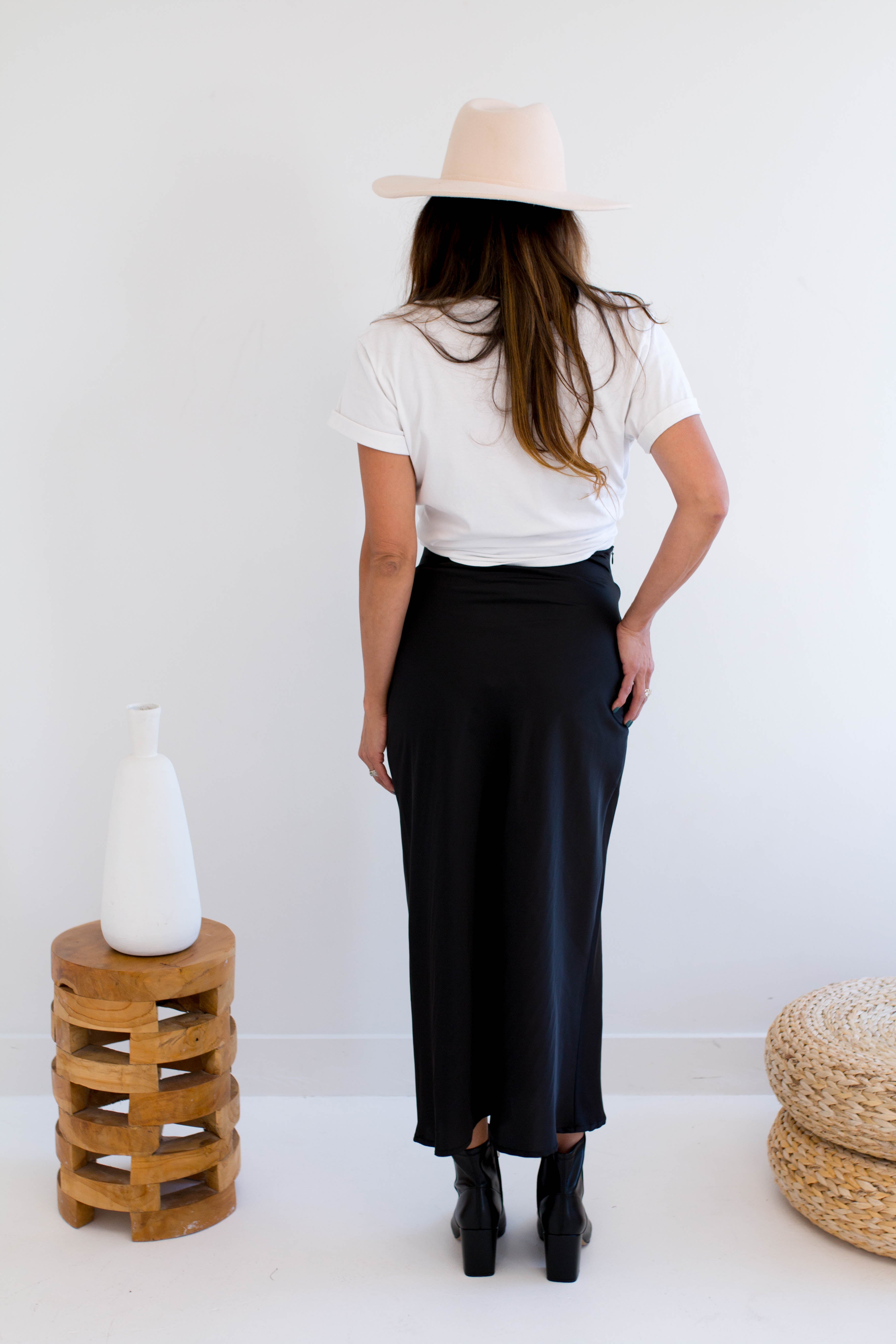 Woman wearing a white t-shirt with text and graphics, black satin maxi skirt, and beige hat in a minimalistic setting. Made in Canada Back View.