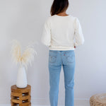 Back view of a woman wearing a cream sweater and light blue jeans standing in a minimalistic room with a wooden stool and decorative items. Made in canada clothing.