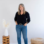 Woman in black bamboo top and blue jeans standing in a minimalistic room with a white wall and wooden furniture.