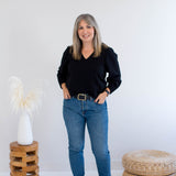 Woman in black bamboo top and blue jeans standing in a minimalistic room with a white wall and wooden furniture.