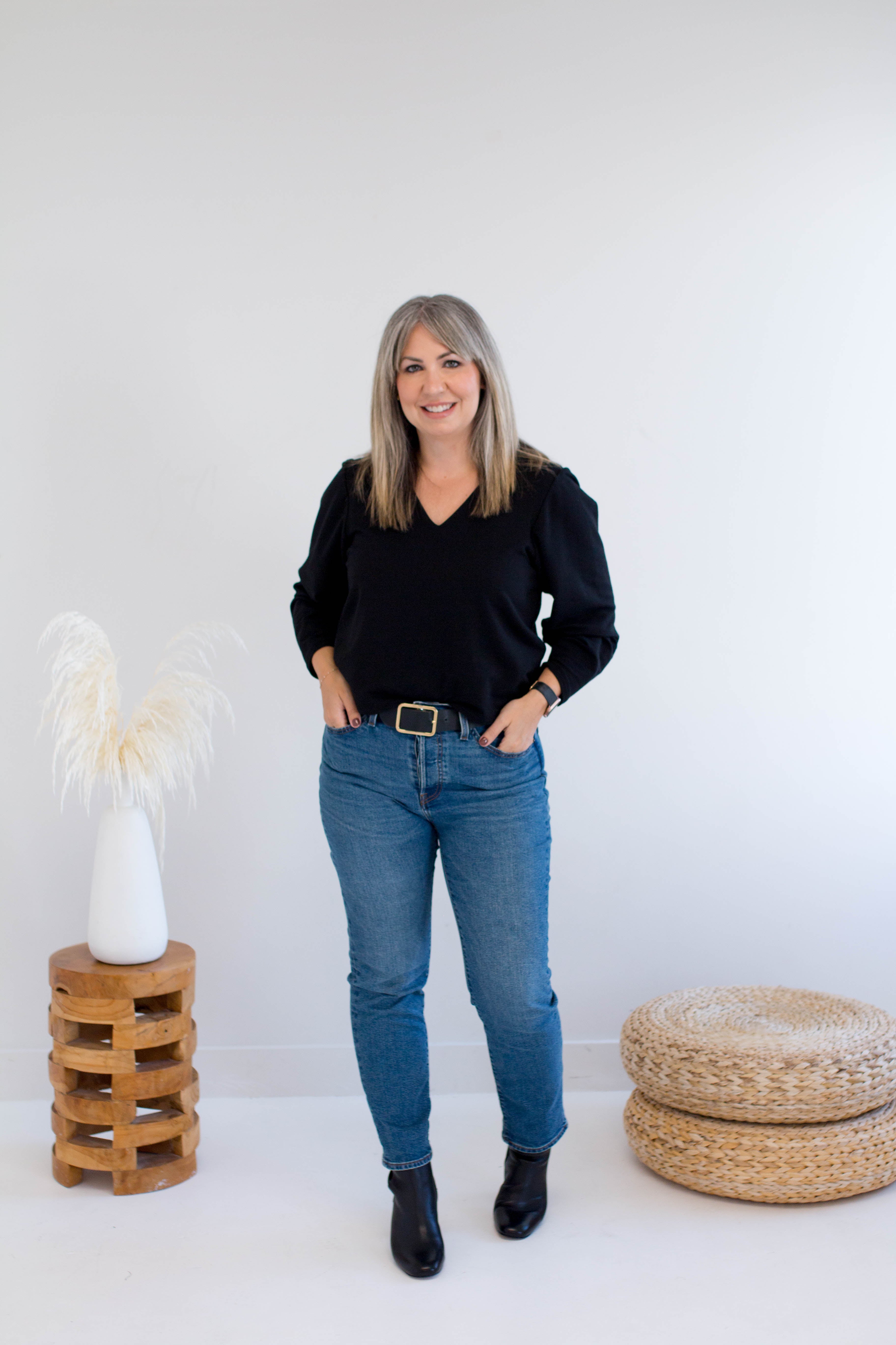 Woman in black bamboo top and blue jeans standing in a minimalistic room with a white wall and wooden furniture.