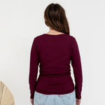 A woman viewed from behind wearing a long-sleeve maroon top and light blue jeans, standing against a plain white wall.