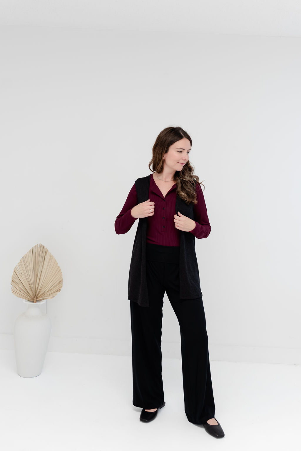 A woman modeling a black sleeveless vest over a burgundy long-sleeve top and black wide-leg pants, standing in a bright white studio with a decorative dried palm in a vase to the left.