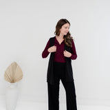 A woman modeling a black sleeveless vest over a burgundy long-sleeve top and black wide-leg pants, standing in a bright white studio with a decorative dried palm in a vase to the left.