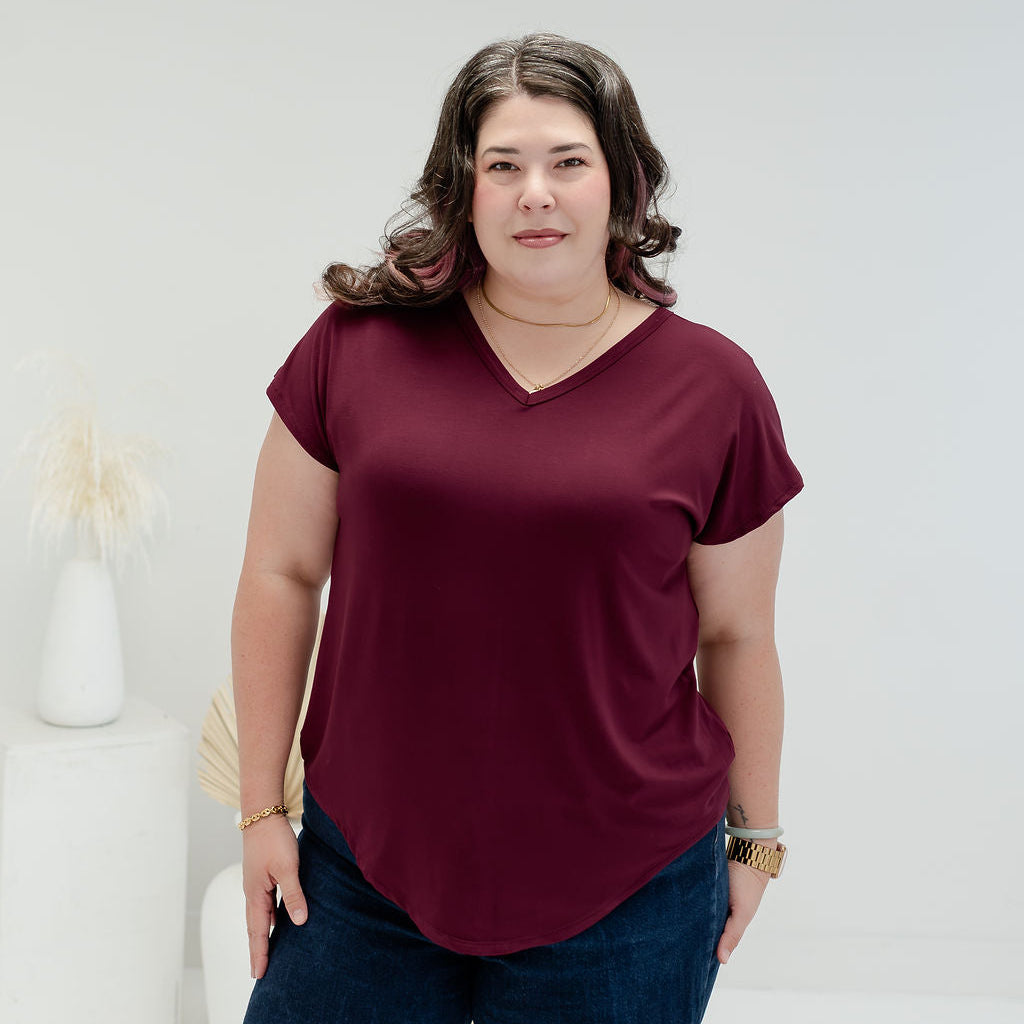 A woman modeling a short-sleeve V-neck top in deep burgundy, paired with dark blue jeans, standing in a bright, minimalist white room. bamboo clothing made in canada