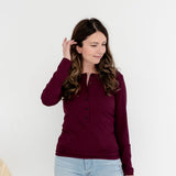 A woman wearing a long-sleeve burgundy henley top and light blue jeans, standing against a plain white studio background.