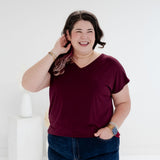 A woman with dark hair smiling and posing in a studio setting, wearing a short-sleeve v-neck burgundy t-shirt and dark blue jeans.  bamboo clothing made in canada