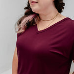 A woman wearing a short-sleeve V-neck t-shirt in deep burgundy, shown from the chest up against a light gray studio background.