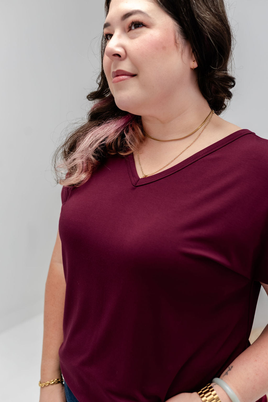 A woman wearing a short-sleeve V-neck t-shirt in deep burgundy, shown from the chest up against a light gray studio background.