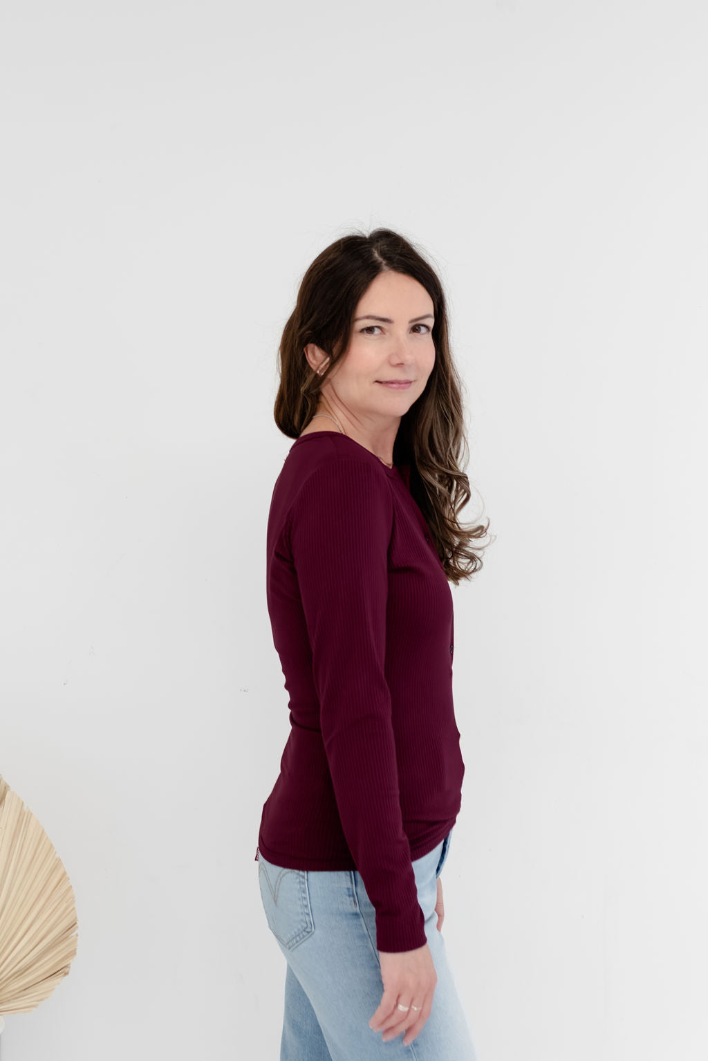 A woman with long brown hair is shown from the side profile, wearing a long-sleeve crewneck top in deep burgundy and light blue jeans, standing against a plain white studio background.