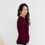 A woman with long brown hair is shown from the side profile, wearing a long-sleeve crewneck top in deep burgundy and light blue jeans, standing against a plain white studio background.