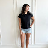 white woman standing against  a white wall wearing a black t-shirt and jean shorts