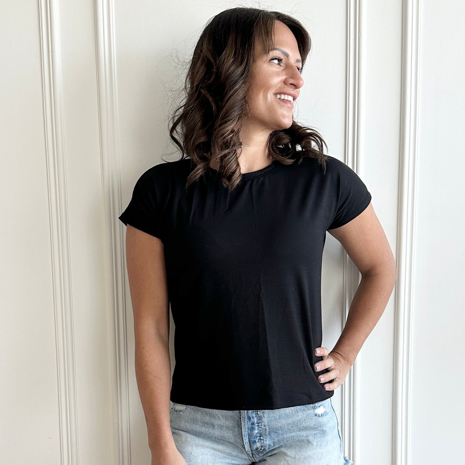 white woman standing against  a white wall wearing a black t-shirt and jean shorts