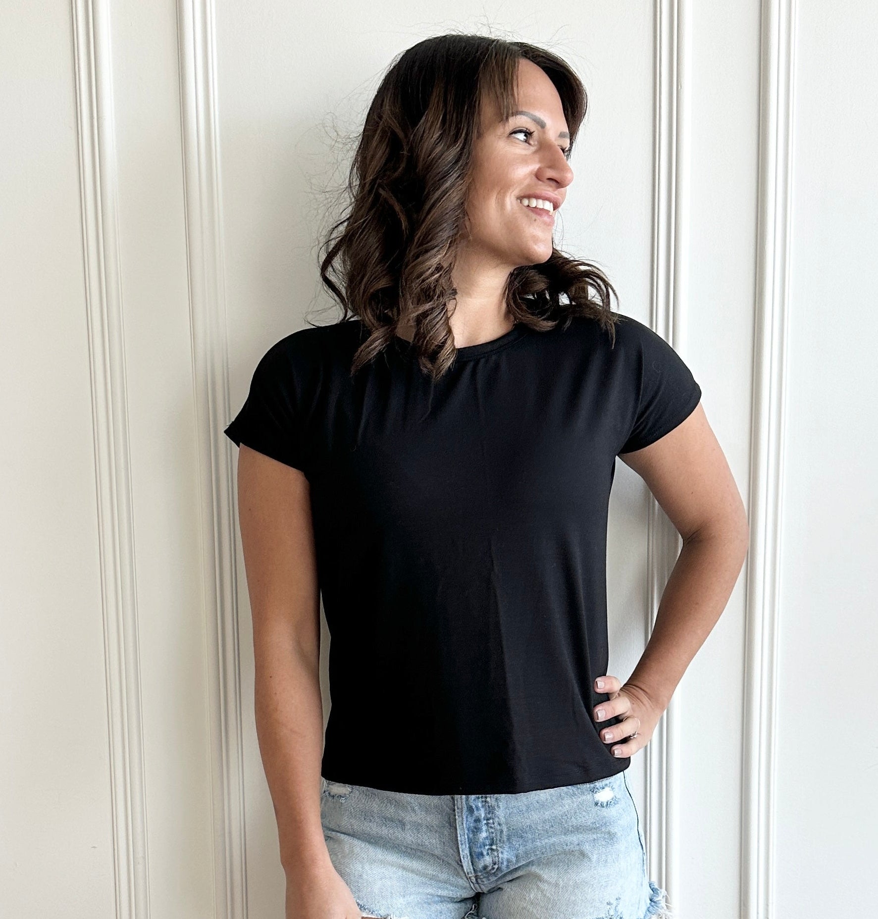 white woman standing against  a white wall wearing a black t-shirt and jean shorts