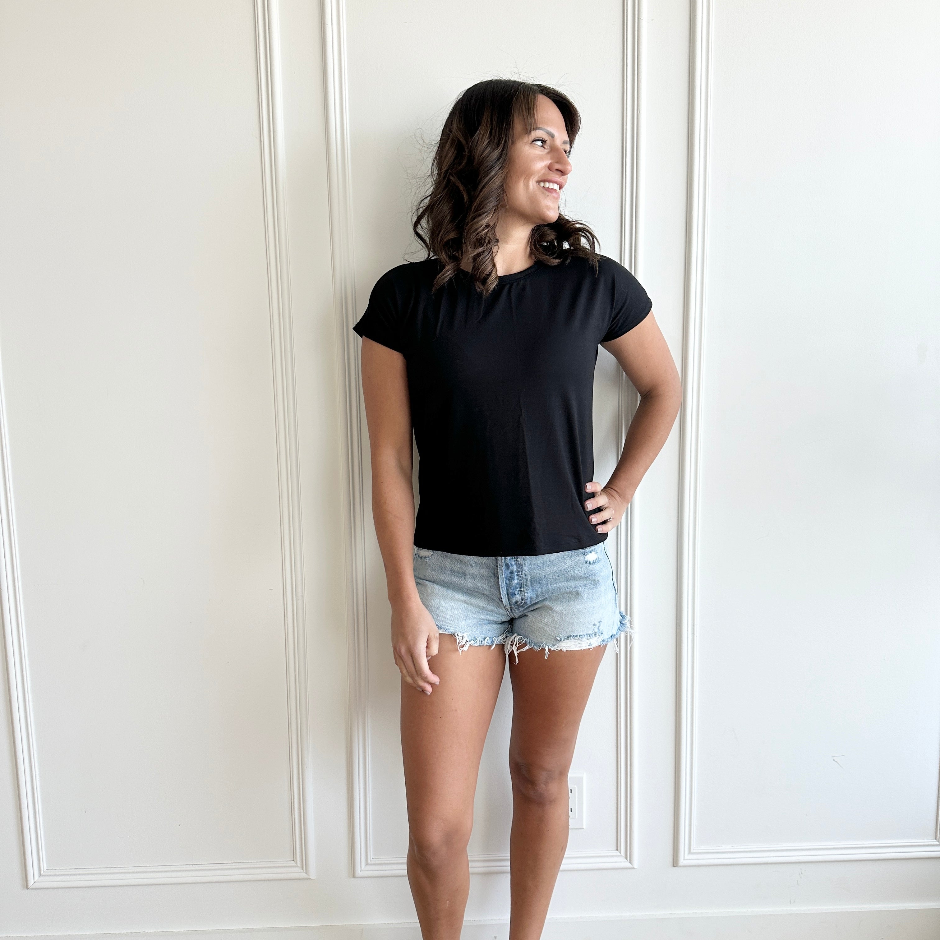 white woman standing against  a white wall wearing a black t-shirt and jean shorts