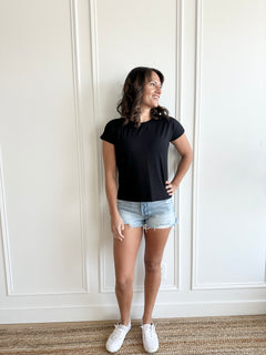 white woman standing against  a white wall wearing a black t-shirt and jean shorts