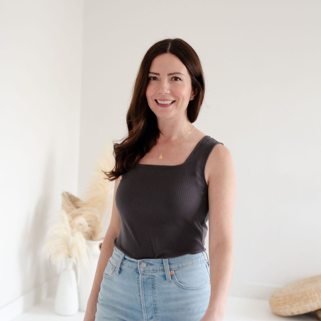 Woman wearing a square neck grey tank top and light blue jeans standing against a white background