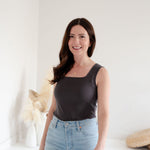 Woman wearing a square neck grey tank top and light blue jeans standing against a white background