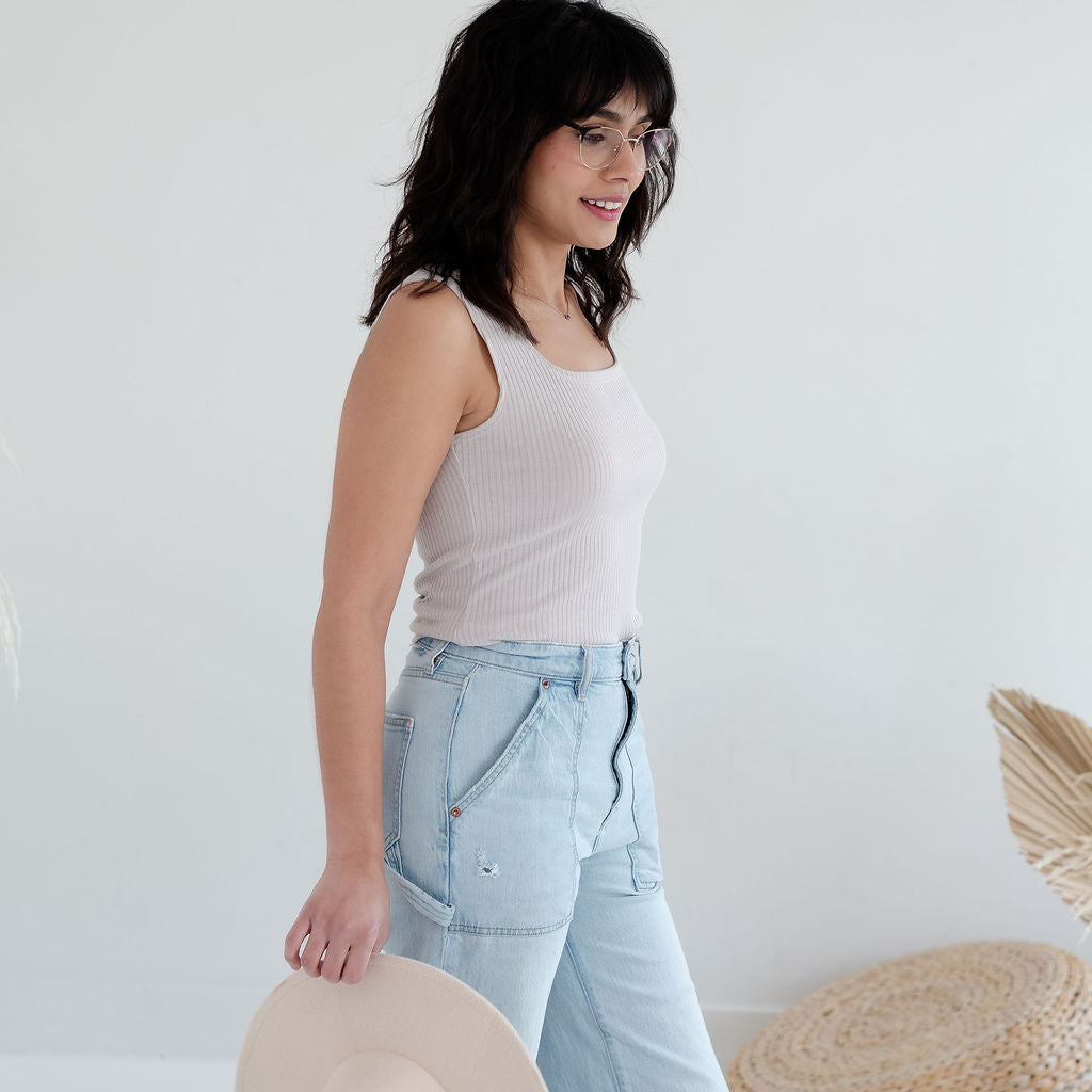 Woman wearing a white tank top and light blue jeans, holding a beige hat against a white background.