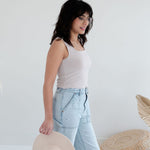 Woman wearing a white tank top and light blue jeans, holding a beige hat against a white background.