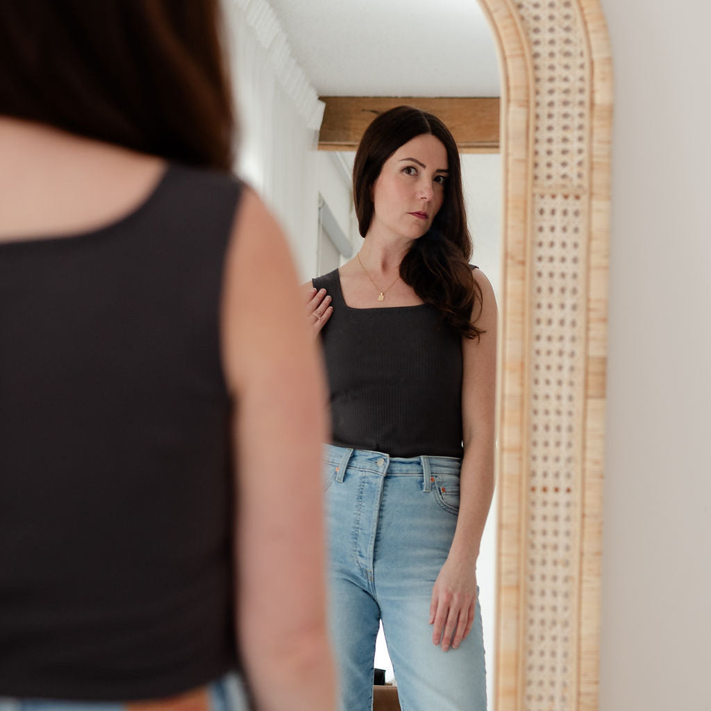 Woman wearing a black tank top and light blue jeans standing in front of a mirror.