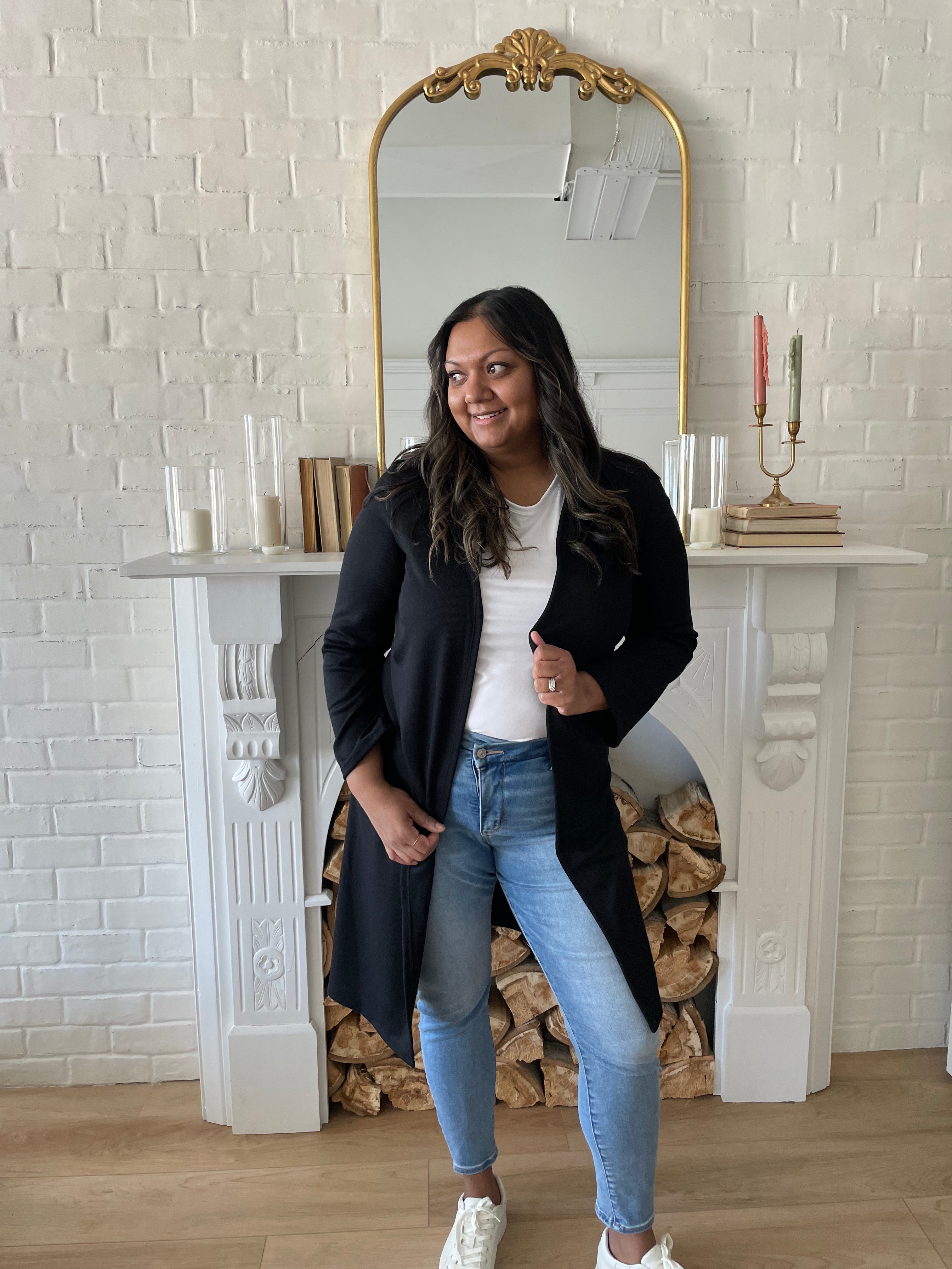 woman wearing a black long duster cardigan and a white crewneck t-shirt with jeans and white sneakers. Standing in front of a mirror and fireplace