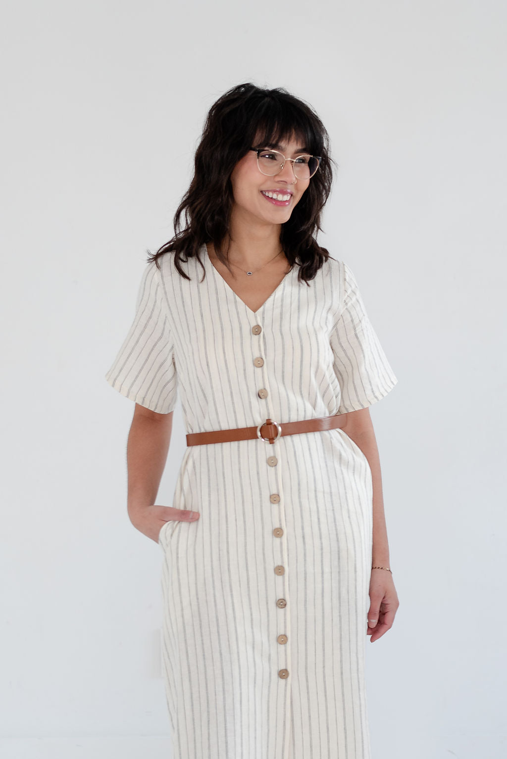 Woman wearing a white dress with buttons and a brown belt on a plain background