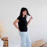 Woman wearing a black t-shirt and light blue jeans standing in a minimalistic room with a white background. Apparel made in Canada