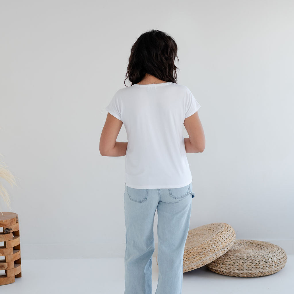 Person wearing a white t-shirt and light blue jeans standing in a minimalistic room with a white wall and wooden. Apparel made in Canadafloor.