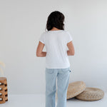 Person wearing a white t-shirt and light blue jeans standing in a minimalistic room with a white wall and wooden. Apparel made in Canadafloor.