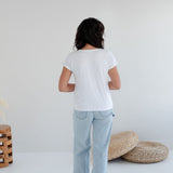 Person wearing a white t-shirt and light blue jeans standing in a minimalistic room with a white wall and wooden. Apparel made in Canadafloor.