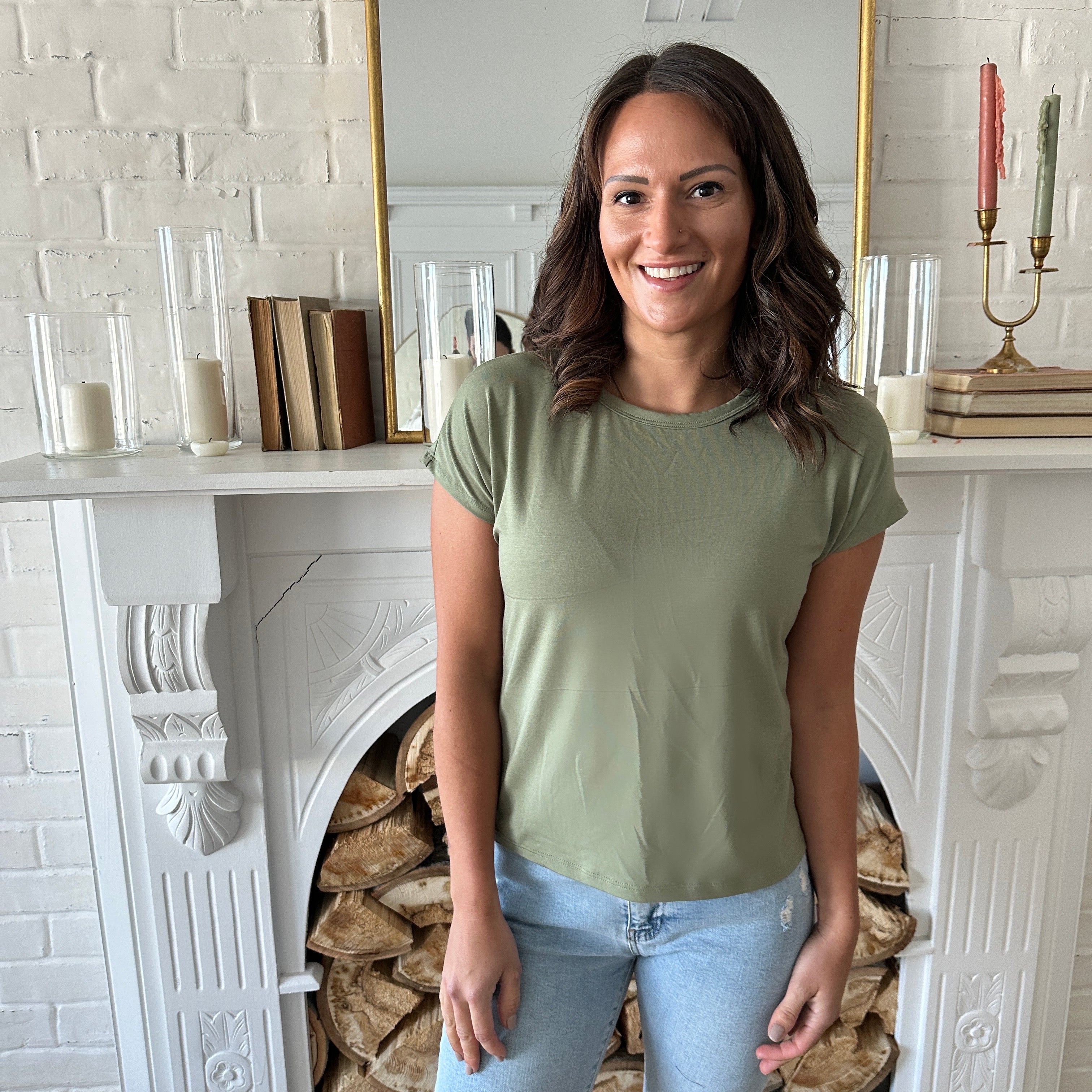 woman smiling standing wearing a sage green bamboo t-shirt and jeans. Standing in front of a fireplace.