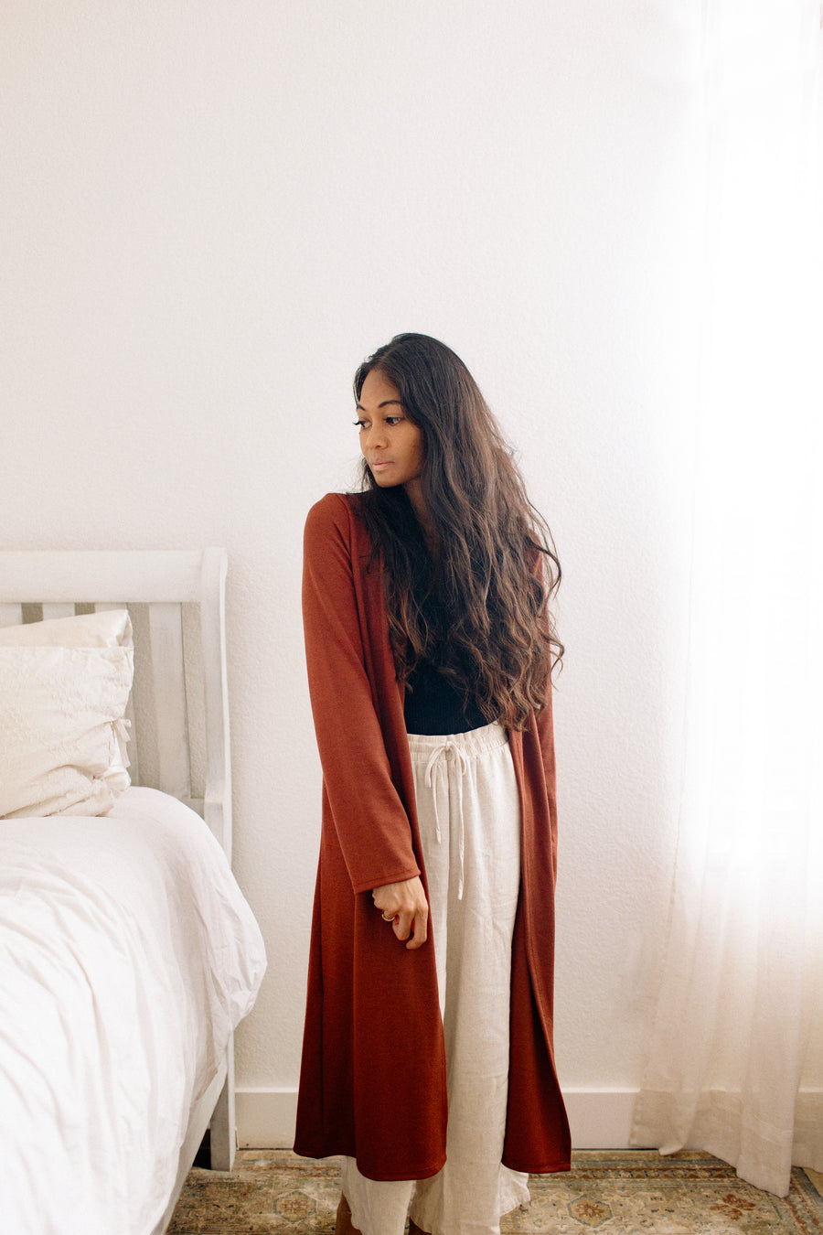 Woman standing in a bedroom with a long red brown cardigan and loungewear