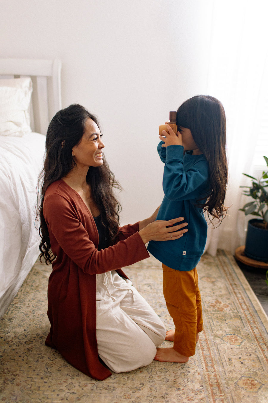 woman wearing a long cardigan and loungewear sitting on the floor with a young girl who is holding a camera
