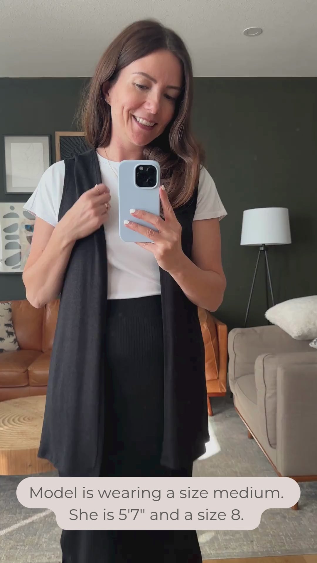 Woman taking a mirror selfie in a living room wearing a black vest and black midi skirt. Made in Canada video.