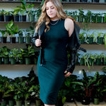 blonde woman wearing emerald green tank dress with a black motorcycle jacket standing in front of a wall of plants