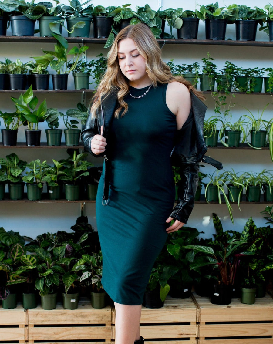 blonde woman wearing emerald green tank dress with a black motorcycle jacket standing in front of a wall of plants