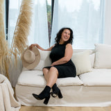 woman smiling with hat in her hand, sitting on a couch wearing a black midi tank dress