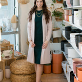 brunette woman wearing a green tank dress with a light pink long cardigan over top. Gold necklaces and tan shoes. standing in front of plant pots with basket lights.