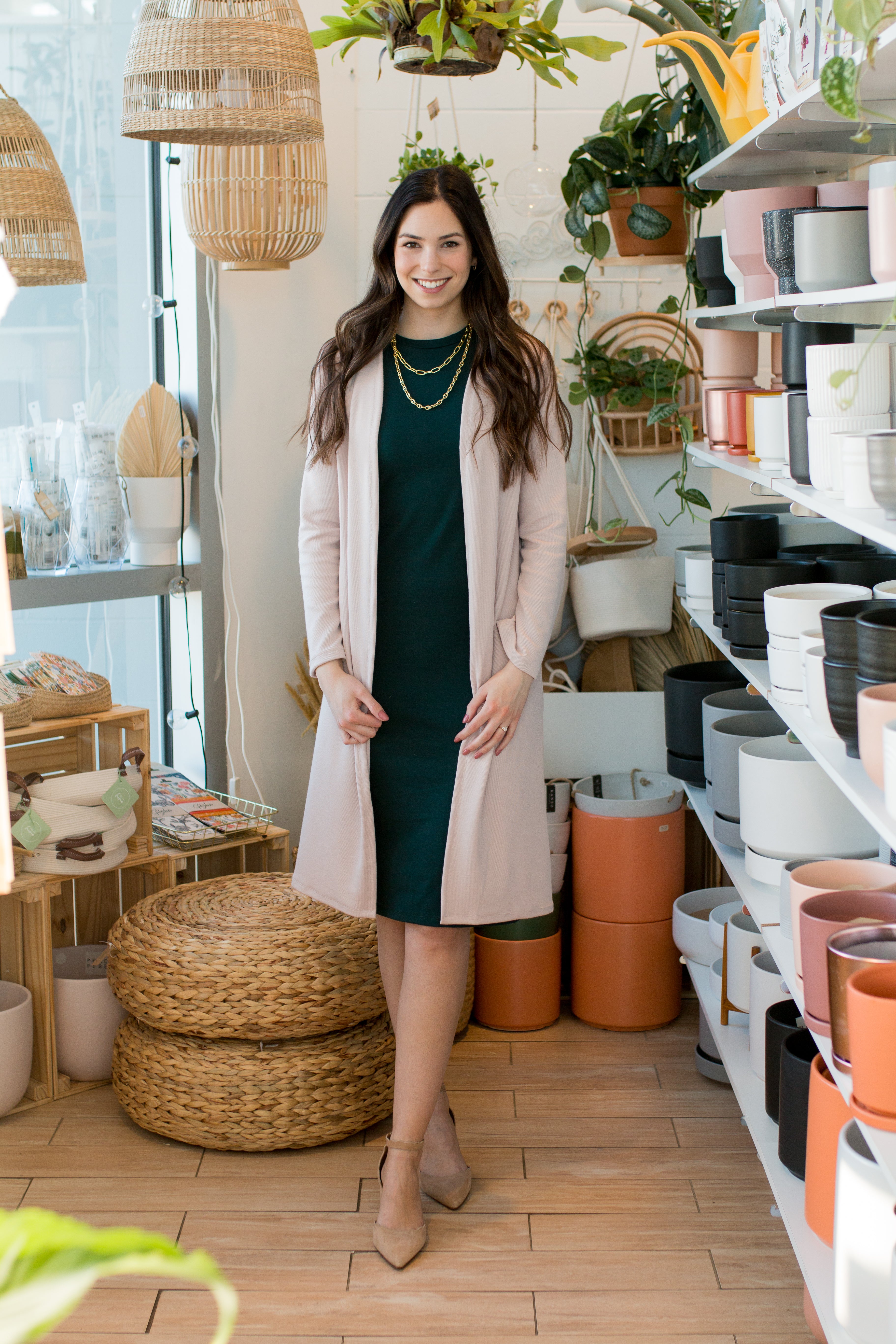 brunette woman wearing a green tank dress with a light pink long cardigan over top. Gold necklaces and tan shoes. standing in front of plant pots with basket lights.