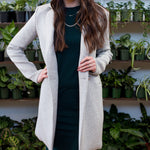 brunette woman wearing a green tank dress, a grey blazer, tan hat and a silver necklace in front of a wall of plants