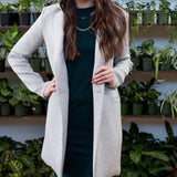 brunette woman wearing a green tank dress, a grey blazer, tan hat and a silver necklace in front of a wall of plants