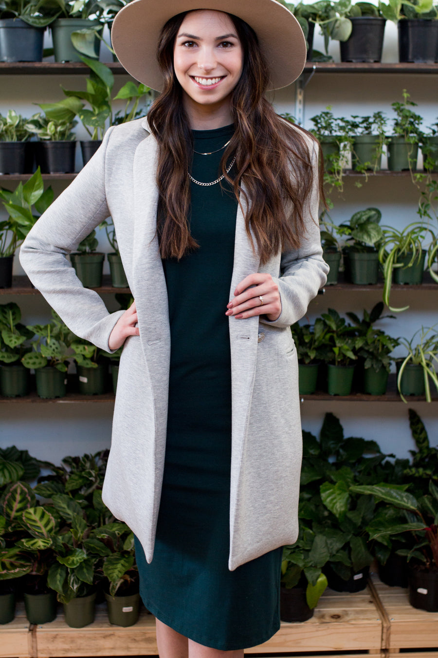 brunette woman wearing a green tank dress, a grey blazer, tan hat and a silver necklace in front of a wall of plants