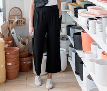 Brunette woman wearing a white t shirt and black wide leg pants standing in a room with plant pots