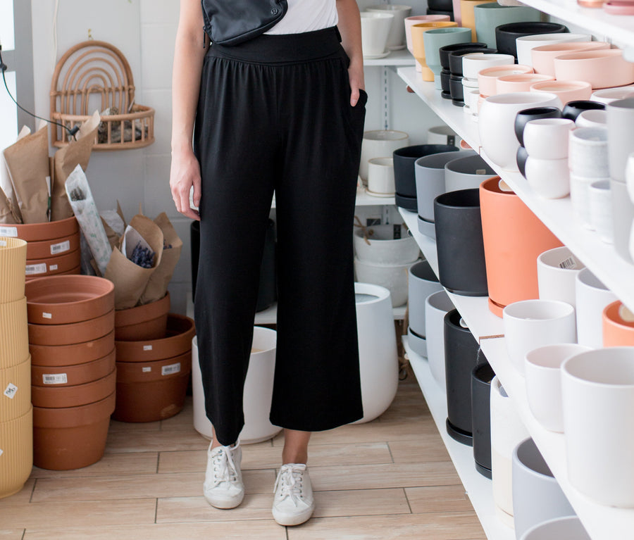 Brunette woman wearing a white t shirt and black wide leg pants standing in a room with plant pots